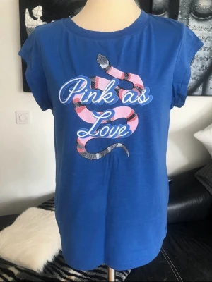 Blå t-shirt med tryck från Coster Copenhagen  - Blå t-shirt med korta ärmar och rund hals. Framsidan har ett stort tryck med en rosa och svart orm samt texten 'Pink as Love'. Skön passform och tillverkad i mjuk bomull, perfekt för en avslappnad stil.