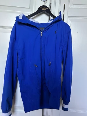 Blå vindjacka från Polo Ralph Lauren - Snygg blå vind jacka från polo! Jackan har två fickor vid magen med dragkedja.