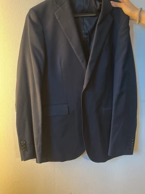 Mörkblå klassisk kavaj - Snygg mörkblå kavaj med två knappar och klassiska slag. Kavajen har två fickor framtill med lock och knappar vid ärmsluten. Perfekt för en stilren och uppklädd look. Materialet ser ut att vara en slät textil, troligen polyester eller ullblandning. Stl 52