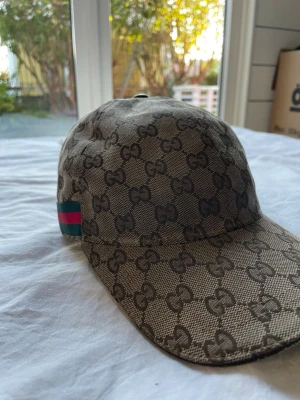 Gucci beige keps med GG-mönster - Säljer en beige keps från Gucci med det klassiska GG-monogrammet över hela kepsen. På sidan finns en grön och röd rand som ger en ikonisk touch. Kepsen har böjd skärm och är tillverkad i ett slitstarkt tygmaterial. Perfekt för dig som vill ha en stilren och lyxig accessoar.