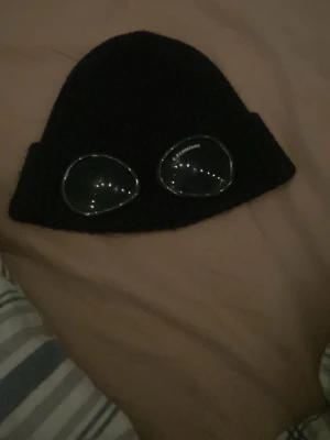 Svart C.P. Company goggle mössa - Säljer en svart ribbstickad mössa från C.P. Company med de ikoniska goggle-linserna framtill. Mössan är i mjukt material och har en cool streetwear-vibe som sticker ut. Perfekt för dig som gillar unika accessoarer och vill ha något extra till din outfit.