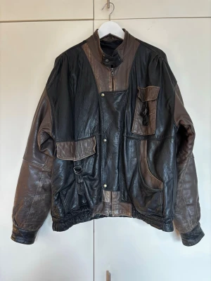 Oversized vintage skinnjacka storlek 52 - Unik oversized skinnjacka med svarta och bruna partier. Jackan har flera stora fickor framtill, ståkrage med spänne och elastisk nederkant. Perfekt för dig som vill sticka ut med en cool vintage-look. Storlek 52/ XL