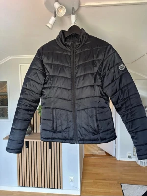 Svart pufferjacka från Peak Performance - Säljer en svart pufferjacka från Peak Performance med diskret logotyp på ärmen. Jackan har hög krage, dragkedja framtill och quiltad design. Perfekt för kalla dagar och riktigt skön att bära. Materialet är syntet och jackan har en klassisk, rak passform.