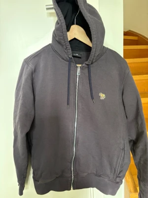 Blågrå hoodie med dragkedja - Paul Smith - Snygg mörkgrå hoodie från Paul Smith med dragkedja framtill och huva med dragsko. Liten broderad zebra-logga på bröstet. Mjuk bomullskänsla och två fickor framtill.
