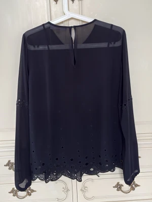 Svart transparent blus från Michael Kors - Stilren svart blus från Michael Kors i transparent material med långärmad design. Blusen har dekorativa håldetaljer längs ärmar och nederkant samt en liten knapp i nacken. Ett transparent svart linne medföljer för att kunna ha unders Perfekt för en trendig och elegant look. Använd 1 gång.