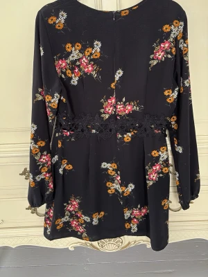 Svart blommig kortklänning med spets - Svart kortklänning med färgglatt blommönster. Klänningen har långa ärmar och en dekorativ spetsdetalj i midjan som ger en unik touch. Perfekt för dig som gillar romantiska och feminina plagg. Storlek 12/38-40