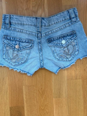 Blå jeansshorts med slitningar och broderade  knappfickor  - Säljer ett par ljusblå jeansshorts med råa kanter och slitna detaljer framtill. Shortsen har klassisk femficksdesign, knapp och dragkedja i midjan. Perfekta för varma dagar och en avslappnad stil.