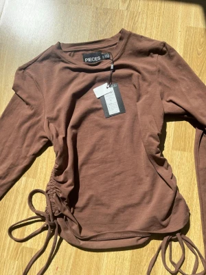 Brun långärmad topp från Pieces - Snygg brun långärmad topp från Pieces med trendig snörning i sidan. Tillverkad i mjuk bomull som känns skön mot huden. Perfekt för dig som gillar en enkel men ändå unik look med coola detaljer.