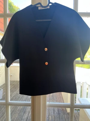Svart kortärmad blus från Zara - Stilren svart blus från Zara med v-ringning och två dekorativa knappar framtill. Blusen har en rak passform och korta ärmar, vilket ger en modern och avslappnad look. Tillverkad i ett mjukt tyg som känns bekvämt mot huden.