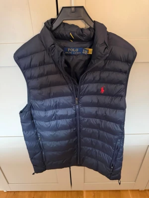 Mörkblå dunväst från Polo Ralph Lauren - Snygg mörkblå dunväst från Polo Ralph Lauren med klassisk quiltad design och dragkedja framtill. Västen har två fickor med dragkedja och ikonisk röd logga på bröstet. Perfekt för lager-på-lager och håller dig varm under kyliga dagar.