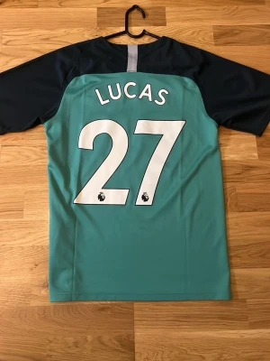 Tottenham Hotspur tröja. 18/19 Lucas moura ikoniska tröja - Snygg replika Tottenham Hotspur bortatröja från Nike med tryck 'LUCAS 27' på ryggen. Tröjan är turkos med mörkblå ärmar och har klubbmärke och sponsorlogga på bröstet. Tillverkad i lätt och ventilerande material, perfekt för fotbollsträning eller match.