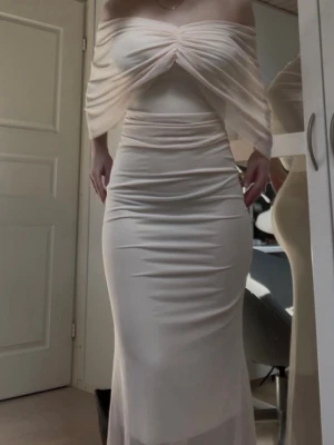 Lång klänning  - Elegant långklänning i champagnefärgat tyg med offshoulder-design och draperad överdel. Klänningen har figurnära passform och är tillverkad i ett glansigt, tunt material som ger en lyxig känsla. Perfekt för speciella tillfällen.