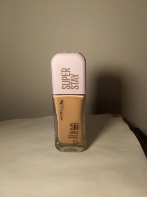 Maybelline Super Stay Foundation 128 - Maybelline Super Stay 30H foundation i nyansen 128. Kommer i en rektangulär glasflaska med ljuslila plastlock. Ger en matt finish och lång hållbarhet. Perfekt för dig som vill ha en jämn och täckande bas.