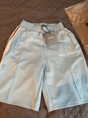 Ljusblå shorts från Palm Angels - Palm Angels shorts i ljusblått med vita sidoränder och elastisk midja. Klassisk sportig look med logga tryckt på vänster ben. Perfekta för en avslappnad streetwear-stil.