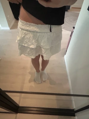 Vit volangkjol med broderi - Söt vit kjol med dubbla volanger och broderade detaljer. Kjolen har resår i midjan för skön passform och är gjord i ett lätt tyg som känns luftigt. Perfekt för sommarens alla tillfällen och enkel att matcha med olika toppar.