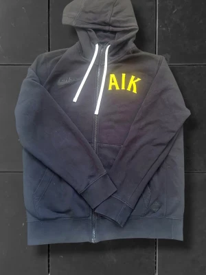 Svart Nike AIK hoodie med dragkedja - Svart hoodie från Nike med AIK-tryck i gult på bröstet. Tröjan har huva med vita snören och dragkedja framtill. Tillverkad i mjukt material, perfekt för en sportig och avslappnad stil. Praktiska fickor på sidorna och klassisk passform. Storlek S