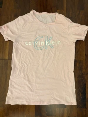 Ljusrosa Calvin Klein t-shirt - Snygg ljusrosa t-shirt från Calvin Klein med tryckt logga i vitt och blått på bröstet. Klassisk passform med korta ärmar och rund halsringning. Perfekt för en avslappnad och trendig stil.