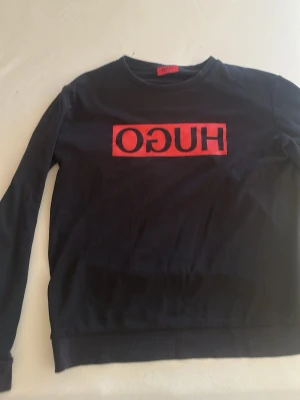 Svart sweatshirt från Hugo Boss - Svart sweatshirt från Hugo Boss med röd logga och spegelvänd text på bröstet. Tröjan har rund halsringning och ribbade muddar vid ärmslut och nederkant. Perfekt för en avslappnad och trendig stil.