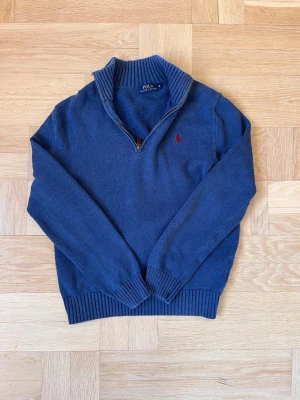 Blå stickad tröja från Polo Ralph Lauren - Mörkblå stickad tröja från Polo Ralph Lauren med halv dragkedja och röd broderad logga på bröstet. Tröjan har ribbstickade muddar och krage, perfekt för lager-på-lager. Klassisk och stilren design.