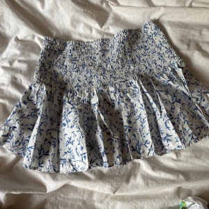 Blåvit blommig volangkjol - Supersöt kort kjol i blått och vitt med blommigt mönster och volang. Kjolen har smock i midjan för en bekväm passform och är gjord i ett lätt bomullstyg. 