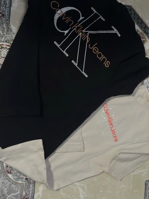 Calvin Klein Jeans t-shirts svart & beige - Två snygga t-shirts från Calvin Klein Jeans, en i svart och en i beige, tillverkade i mjuk bomull. Perfekta för att lyfta din outfit med stilren design och ikoniskt märke.