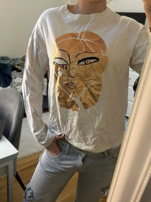 Vit långärmad topp med ansiktsmotiv - Vit långärmad topp med ett stort grafiskt ansiktsmotiv i guld och beige på framsidan. 