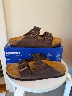 Birkenstock Arizona Mocha sandaler - Birkenstock Arizona sandaler i färgen mocha med två justerbara remmar och metallspännen. Ovandel i mocka och fotbädd i kork med mjuk innersula. Klassisk och bekväm modell som passar perfekt till sommaren.
