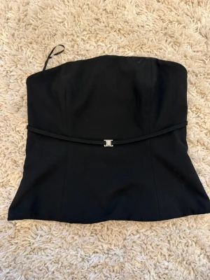 Svart tubtopp från bikbok - Snygg svart tubtopp från Bikbok med smal dekorativ rem och metallspänne framtill. Toppen har en figurnära passform och stängs med två spännen på insidan. Perfekt för en stilren look. Öppen i ryggen