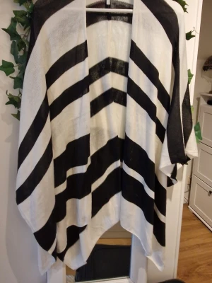 Svartvit poncho med grafiskt mönster - Säljer en oversized poncho i svart och vitt med breda, grafiska ränder i ett V-format mönster. Ponchon har öppen front och faller snyggt över axlarna. Perfekt för lager på lager och ger en cool, avslappnad vibe till din outfit.