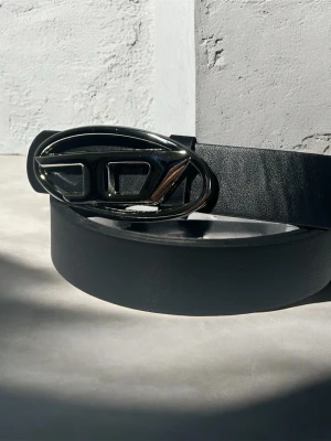 Svart bälte med silverfärgat spänne - Stilrent svart bälte från diesel med ett brett band i skinn och ett unikt, ovalt silverfärgat spänne med logodetalj. Perfekt accessoar för att lyfta din outfit med en modern touch.
