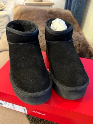 Svarta mockaboots liknar Ugg från s.Oliver 39 - Snygga svarta boots från s.Oliver med mjuk insida och kraftig sula. De har en enkel, stilren design i mocka och är perfekta för kalla dagar. Skafthöjden är låg och de är lätta att dra på. Passar dig som gillar en clean och modern look. Endast testade en dag. 