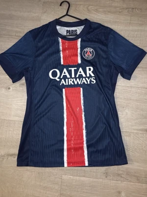 PSG matchtröja blå/röd - Snygg PSG matchtröja i mörkblått med bred röd vertikal rand och vita detaljer. Tröjan har klubbmärke på bröstet och Qatar Airways som sponsortryck. Tillverkad i lätt och ventilerande material, perfekt för fotboll eller träning.