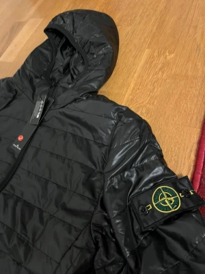 Svart dunjacka från Stone Island - Snygg svart dunjacka från Stone Island med ikonisk patch på ärmen. Jackan har huva, dragkedja framtill och quiltad design. Perfekt för kalla dagar och ger en clean streetwear-look. Materialet är glansigt och lätt, vilket gör den både bekväm och stilren.