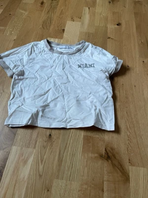 Vit t-shirt med beige detaljer MIAMI - Vit t-shirt från 157 med beige kant runt halsen och ärmsluten. Trycket 'MIAMI' i grått på bröstet ger en clean vibe. Perfekt till jeans eller shorts för en chill look.