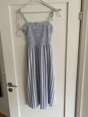 Randig långklänning från H&M Divided - Långklänning från H&M Divided med blå och vita vertikala ränder. Klänningen har smock upptill, tunna axelband med knyt och en luftig passform. Perfekt för varma dagar och har en somrig vibe.