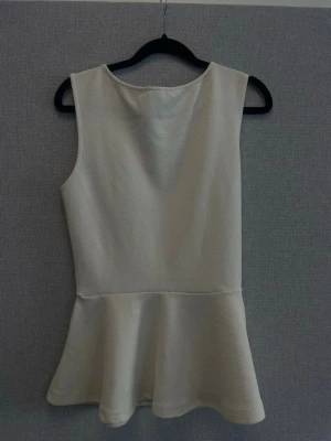 Vit ärmlös kortklänning - Säljer en stilren vit kortklänning med ärmlös design och rund hals. Klänningen har en lätt utsvängd kjol och är tillverkad i ett strukturerat tyg som ger en modern känsla. Perfekt för dig som gillar enkel och fräsch stil.