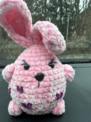 Rosa virkad kanin med lila detaljer - Söt och fluffig virkad kanin i ljusrosa garn med stora öron, svarta ögon och nos samt lila virkade detaljer på kroppen. Kaninen har en rund form och små tassar. Perfekt som dekoration eller gosedjur.