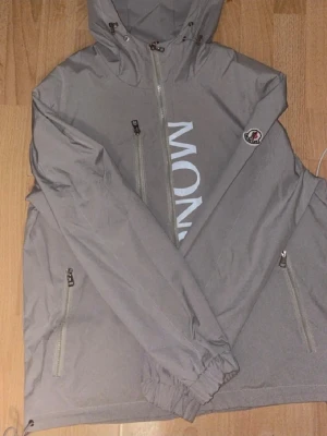 Grå vindjacka från Moncler med huva - En helt ny moncler jacka. Jag säljer den då den är för liten för mig. Den passar en liten M                                                                                Snygg grå vindjacka från Moncler med stor logga framtill, huva med dragsko och dragkedjor på fickorna. Jackan har långa ärmar, diskret Moncler-märke på ärmen och är tillverkad i lätt polyester. Perfekt för dig som gillar stilren och sportig look.