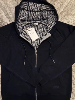 Svart Dior hoodie med monogramhuva - Snygg svart hoodie från Dior med dragkedja och huva fodrad i klassiskt Dior-monogram. Hoodien har dragsko i huvan, fickor framtill och diskret Dior-brodyr på bröstet. Perfekt för dig som vill ha en exklusiv och trendig look.