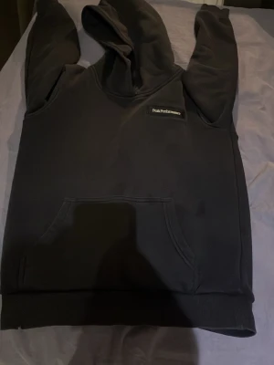 Svart hoodie från Peak Performance - Säljer en svart hoodie från Peak Performance med klassisk logga på bröstet. Hoodien har en stor magficka och ribbade muddar vid ärmslut och nederkant. Perfekt för dig som gillar enkel och stilren design.