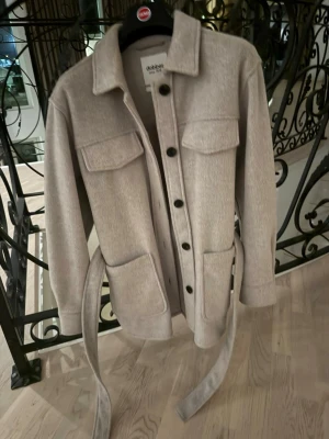 Beige overshirt med bälte från Dobber - Stilren beige overshirt från Dobber med bälte i midjan, stora bröstfickor med lock och knäppning framtill. Tillverkad i ett mjukt tyg som ger en avslappnad men trendig look. Perfekt lager på lager-plagg för dig som gillar minimalistisk stil.