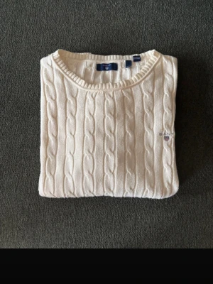 Vit kabelstickad tröja från Gant - Kabelstickad tröja i offwhite från Gant med rund halsringning och diskret logga broderad på bröstet. Tröjan har långa ärmar och ribbade muddar vid ärmslut och nederkant. Perfekt för en klassisk och stilren look.