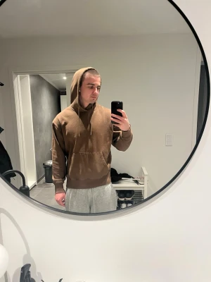 Brun hoodie med huva och magficka - Säljer en enkel brun hoodie med huva och dragsnören. Hoodien har en stor magficka framtill och ribbade muddar vid ärmslut och nederkant. Perfekt för chill dagar eller när du vill ha en avslappnad stil. Materialet känns mjukt och bekvämt.