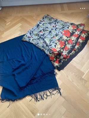 Två sjalar, blommig & marinblå - Säljer två snygga sjalar. En är marinblå med fransar i kanterna, och den andra har ett färgstarkt blommönster i rött, beige, grönt och svart. Båda är tunna och mjuka i materialet.