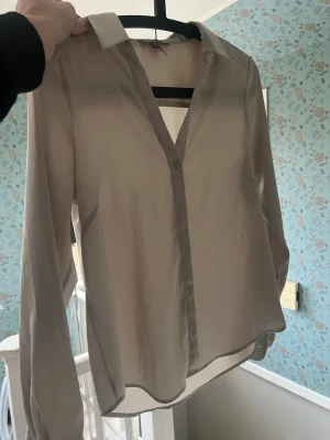 Beige transparent blus med krage - Stilren beige blus i transparent material med klassisk krage och knappdetalj framtill. Långärmad modell med lätt rundad nederkant och breda manschetter. Perfekt för lager på lager och en clean look. Från hm gott skick 