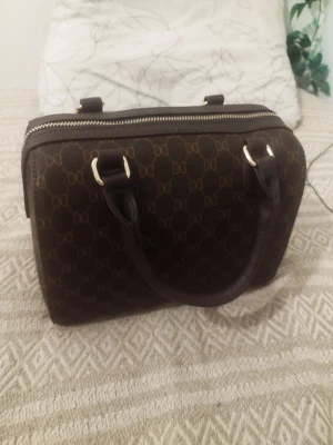 Brun handväska  - Ny pris 500 kr Snygg brun handväska med klassiskt monogrammönster och guldfärgade detaljer. Väskan har två handtag och en avtagbar axelrem för extra flexibilitet. Tillverkad i konstläder med dragkedja upptill och stilren, boxig form.