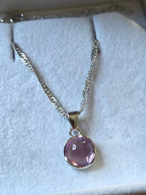 Silverhalsband med lila sten - Snyggt halsband i silverfärg med en rund, lila sten som hänge. Kedjan har en klassisk design och stenen är infattad i en enkel metallram. Perfekt för dig som gillar stilrena smycken med en färgklick.