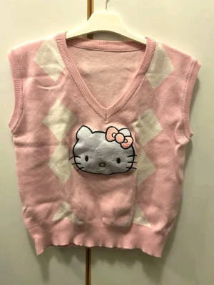 Hello Kitty rosa väst - Söt rosa väst med v-ringning och Hello Kitty-motiv på framsidan. Västen har vita rutor i ett rutigt mönster och är gjord i mjukt stickat material. 