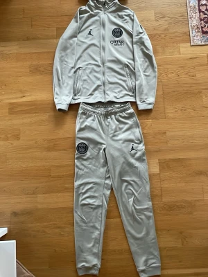 PSG träningsdress - Snygg grå träningsdress från PSG och Jordan med hel dragkedja på jackan och huva. Setet har PSG-logga och Jordan-logga på både jacka och byxa samt Qatar Airways-tryck. Tillverkad i mjukt, stretchigt material som passar perfekt till träning.