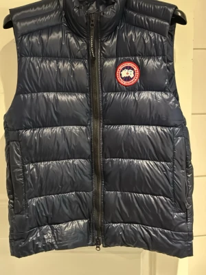 Mörkblå dunväst från Canada Goose - Säljer en mörkblå dunväst från Canada Goose med klassisk logotyp på bröstet. Strl M, men passar även L. Västen har hög krage, dragkedja framtill och quiltad design. Perfekt för lager-på-lager och håller dig varm under kyliga dagar. Köpt från CareOfCarl.  Kan gå ned i pris vid snabb affär! 